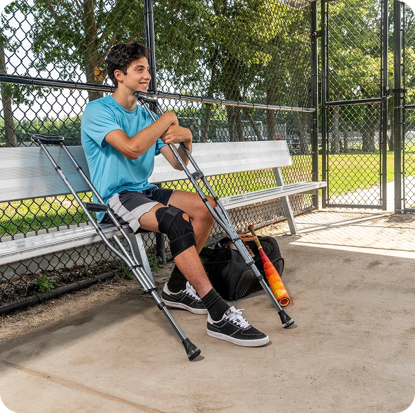 Person with crutches sitting on a bench, knee brace on left leg, baseball bat nearby.