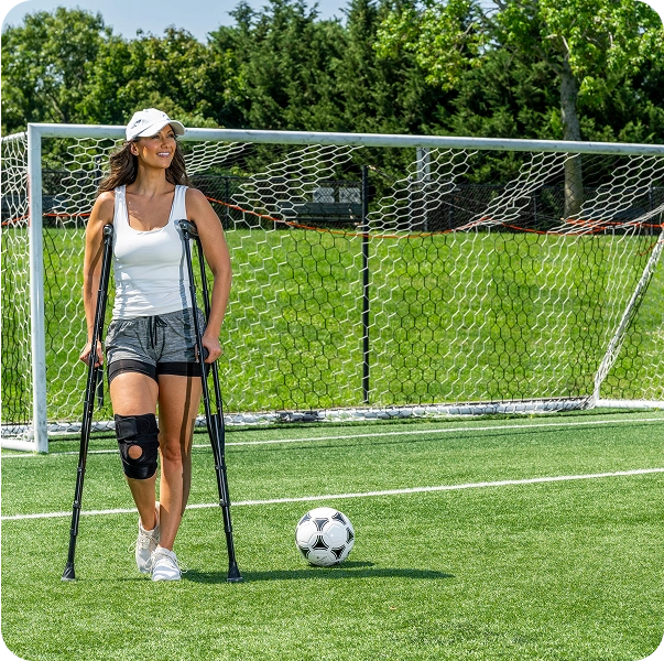 Person with crutches and knee brace on soccer field near ball.