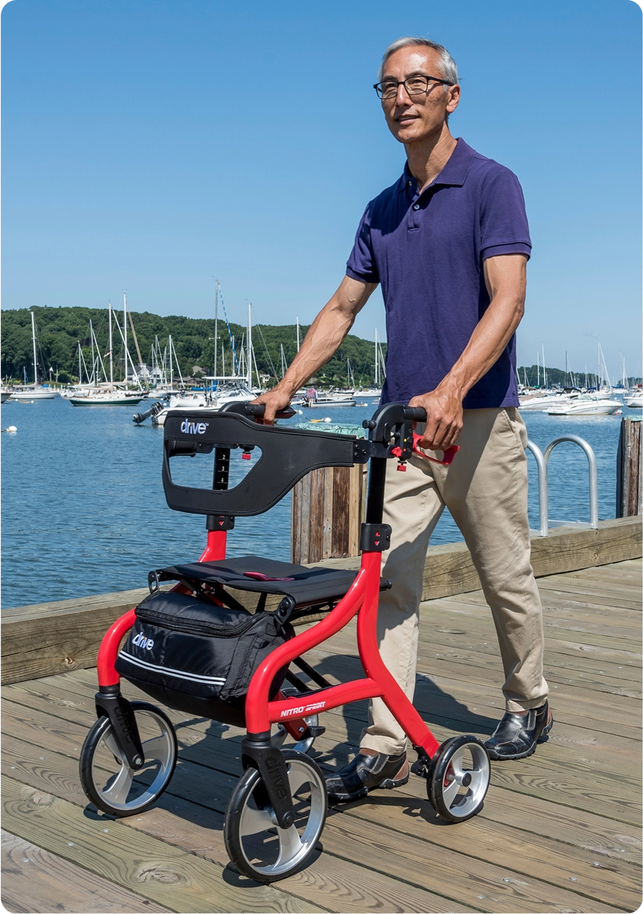 Man walking on a pier with a red walker near boats.