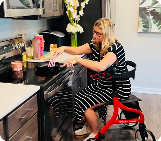 Person in striped dress cooking while sitting on a knee scooter in a kitchen.