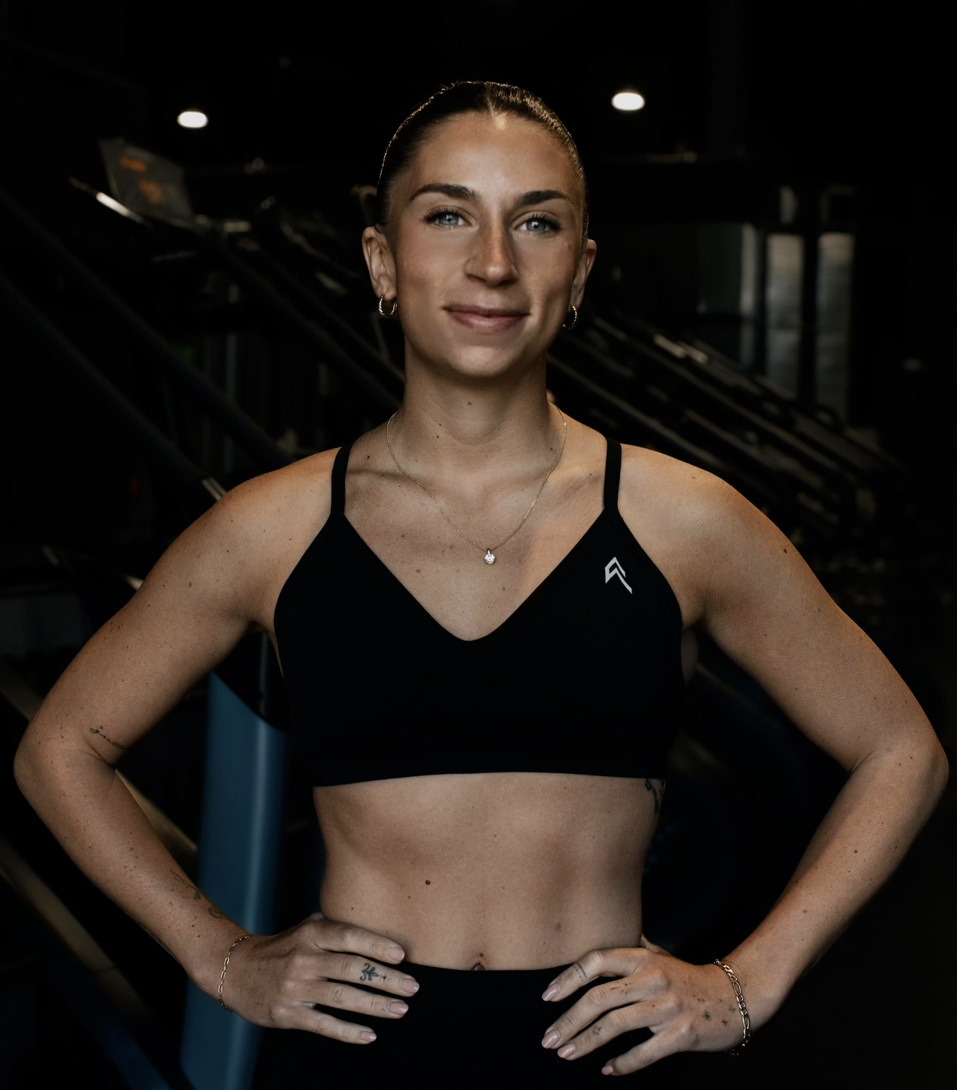 A woman in a black sports bra with her hair pulled back, standing with her hands on her hips.