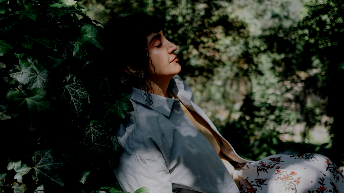 Person in a light shirt resting against green foliage, eyes closed.