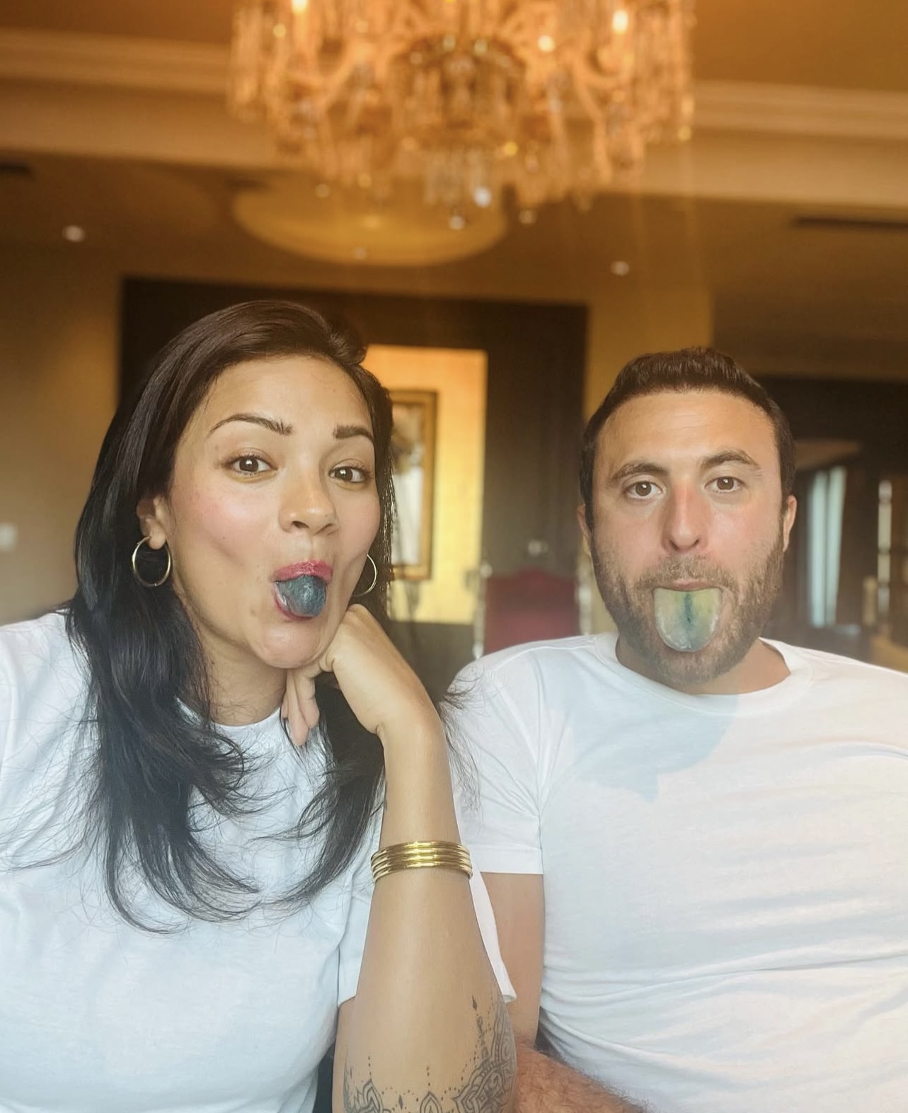 Two people with colored tongues, sitting under a chandelier.