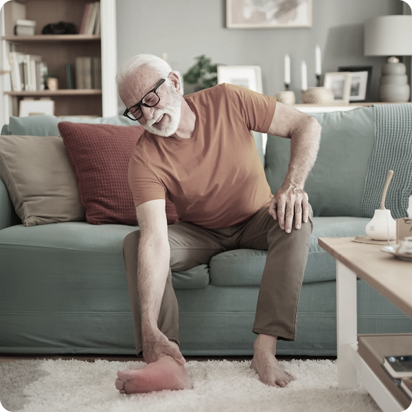An older man sits on a sofa, grimacing in pain while holding his red, swollen foot.