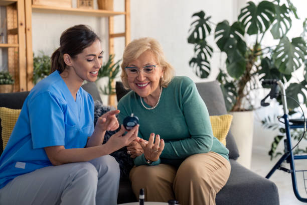 Healthcare worker showing an older woman a medical device, both smiling on a couch.