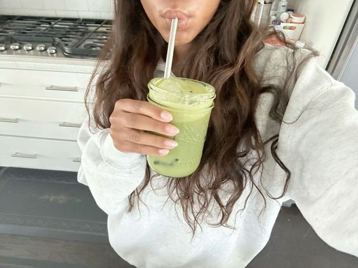 Person drinking green smoothie from a jar with a straw in a kitchen.