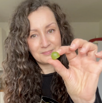 A person with curly hair holding a small green grape close to the camera.