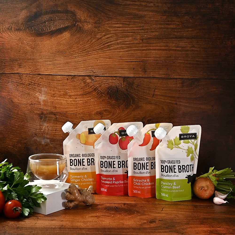 Four bone broth pouches with ingredients and a steaming cup on a wooden surface.