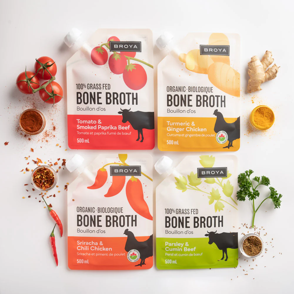 Four Broya bone broth pouches with ingredients like tomatoes, turmeric, and parsley nearby.