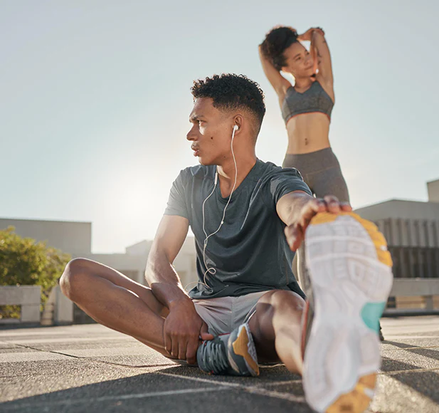 Two people stretching outdoors, one seated listening to music, the other standing with arms raised.