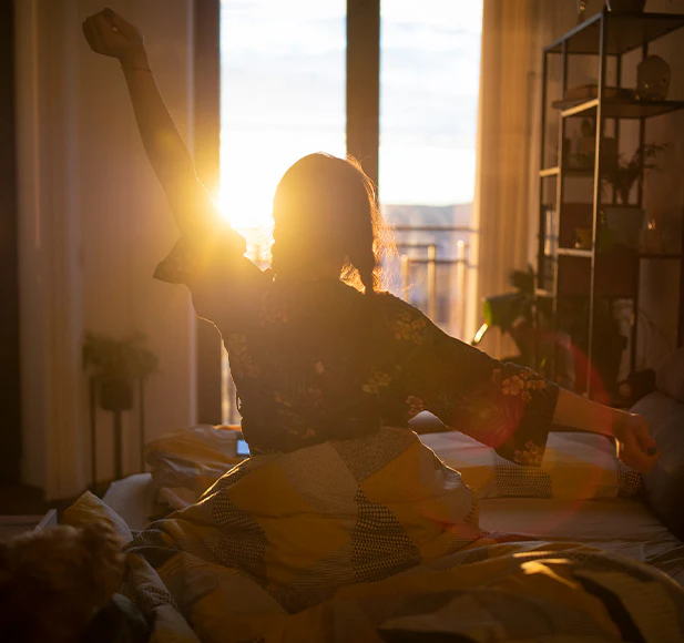 Person stretching in bed with sunrise through window.