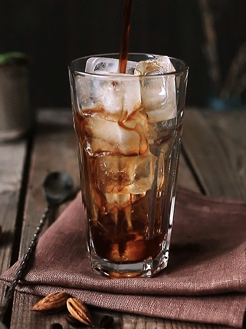 Iced coffee being poured into a glass with ice cubes on a wooden table.