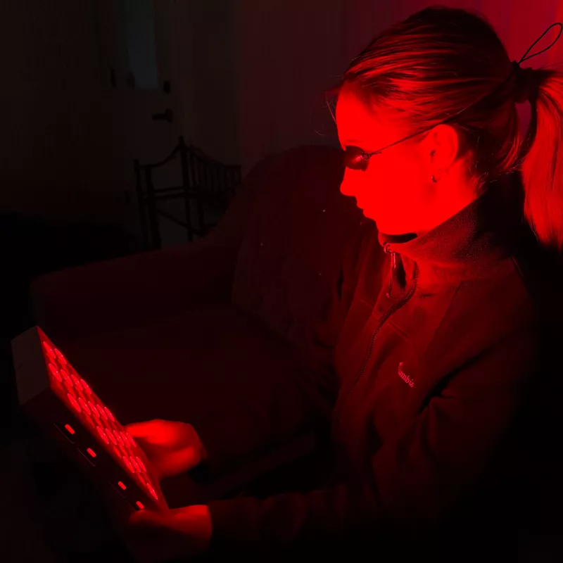 A woman wearing protective eyewear sits in a dark room holding a red light therapy device.