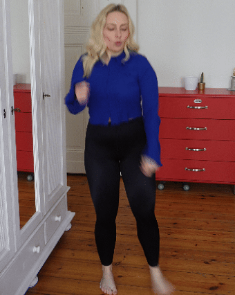 Person standing indoors wearing a blue top and black pants, with a red dresser in the background.