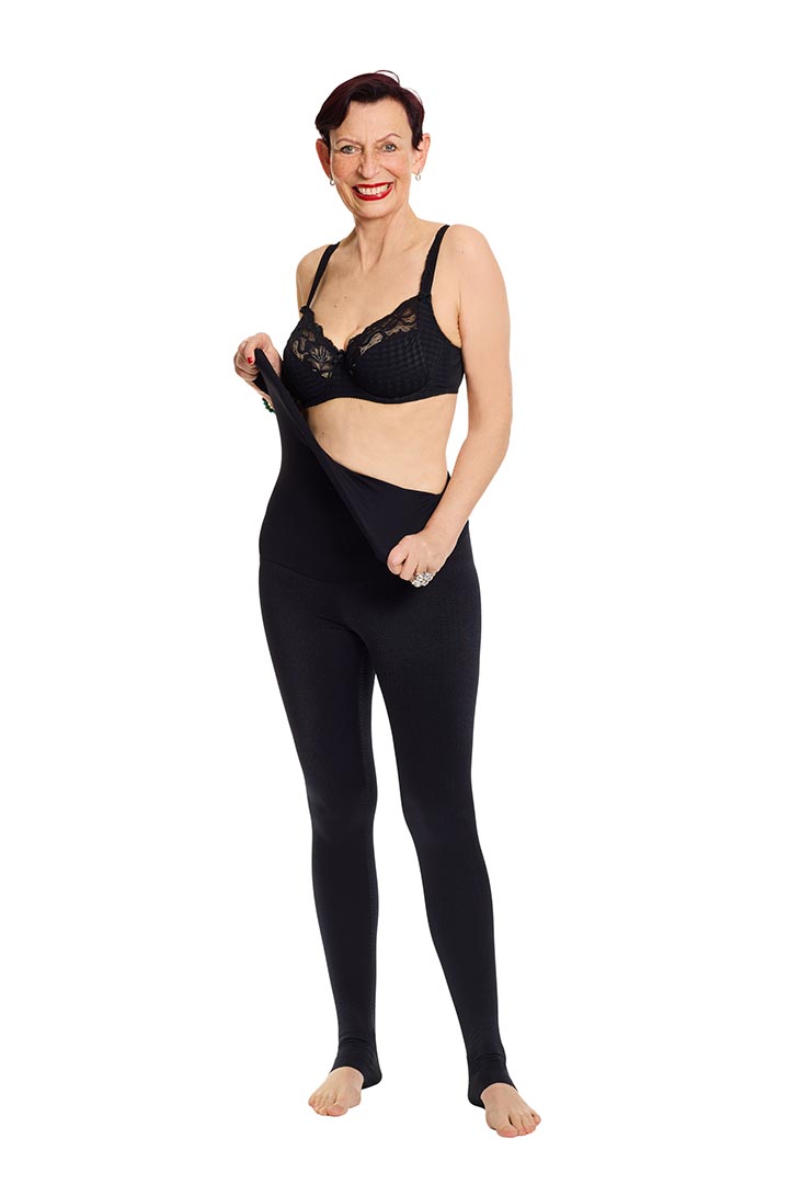 A woman in a black bra stretches the high waistband of her black leggings against a white background.