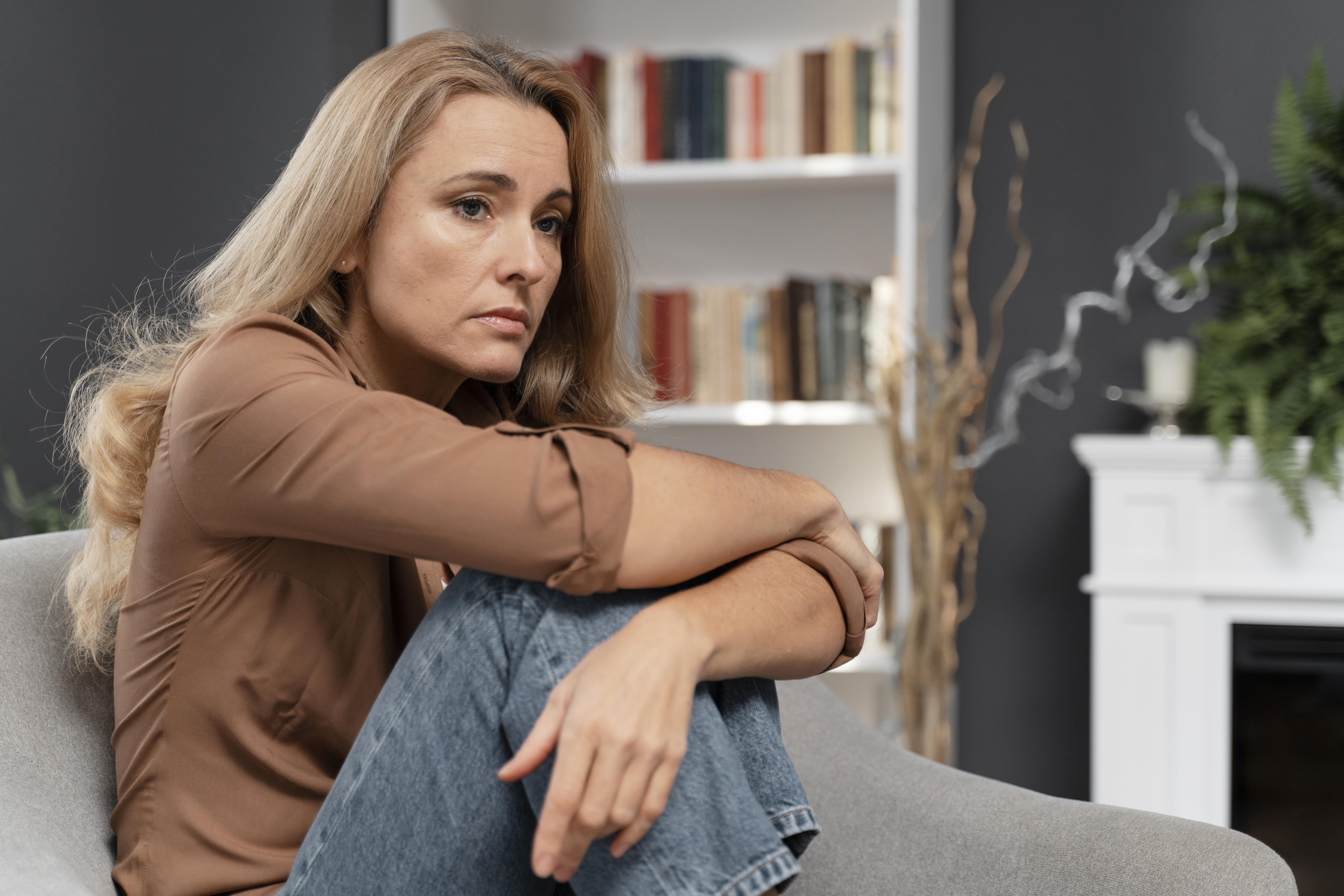 A pensive blonde woman in a brown shirt and jeans sits on a couch, hugging her knees.