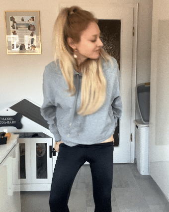 Woman in a grey sweater and black pants standing indoors, looking sideways.