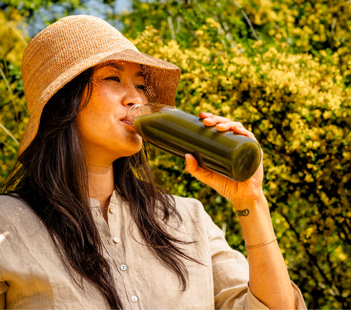 Woman in a sun hat drinking DailyBasis Cycle Routine supplement outdoors surrounded by greenery