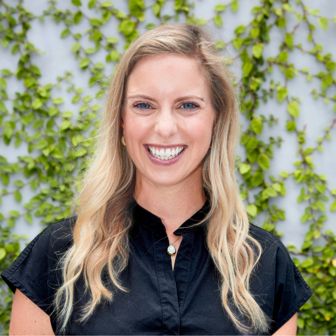 Headshot of a smiling woman with blonde hair, wearing a black shirt, in front of an ivy-covered wall.