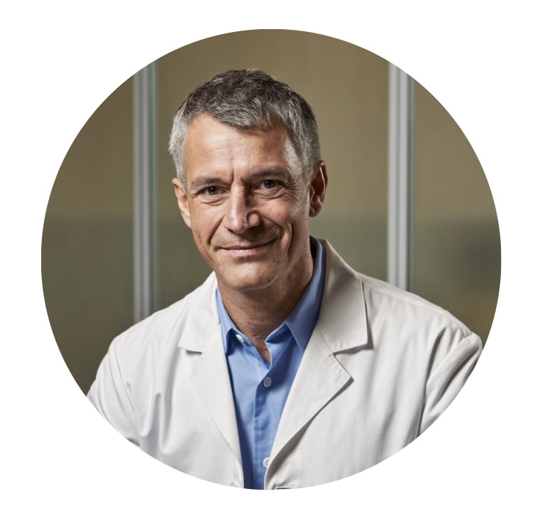 A circular portrait of a smiling middle-aged man with gray hair, wearing a white lab coat.