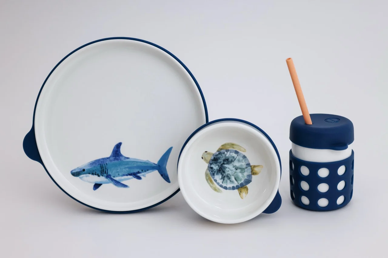 A children's dinnerware set with a shark on the plate, a turtle on the bowl, and a lidded cup.