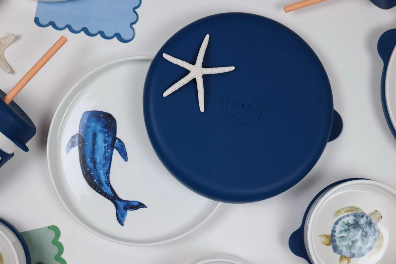 An overhead view of a children's tableware set with a whale plate and a blue lid with a starfish.