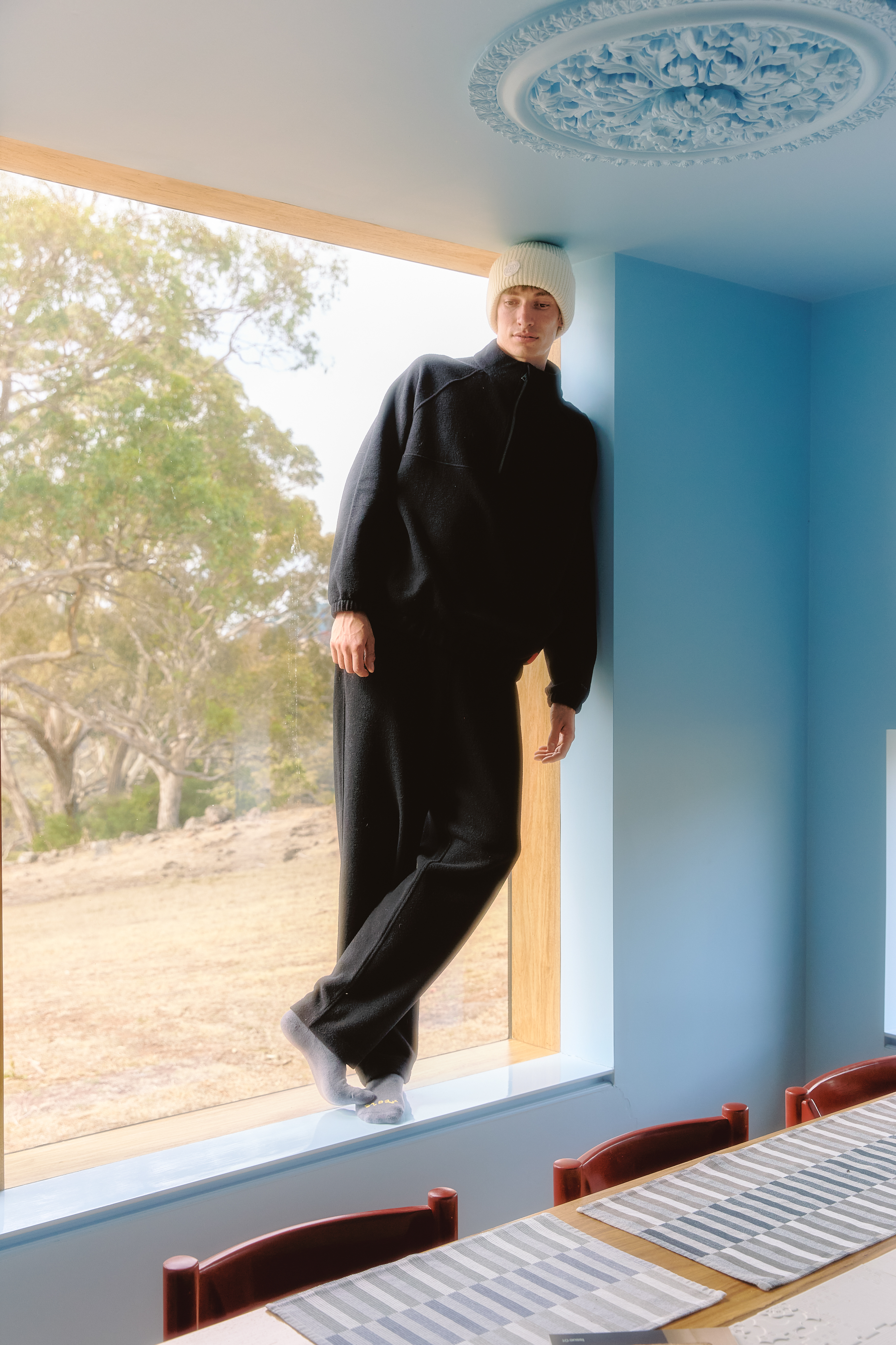 Person leaning in a blue room with a large window, wearing a black outfit and white hat.