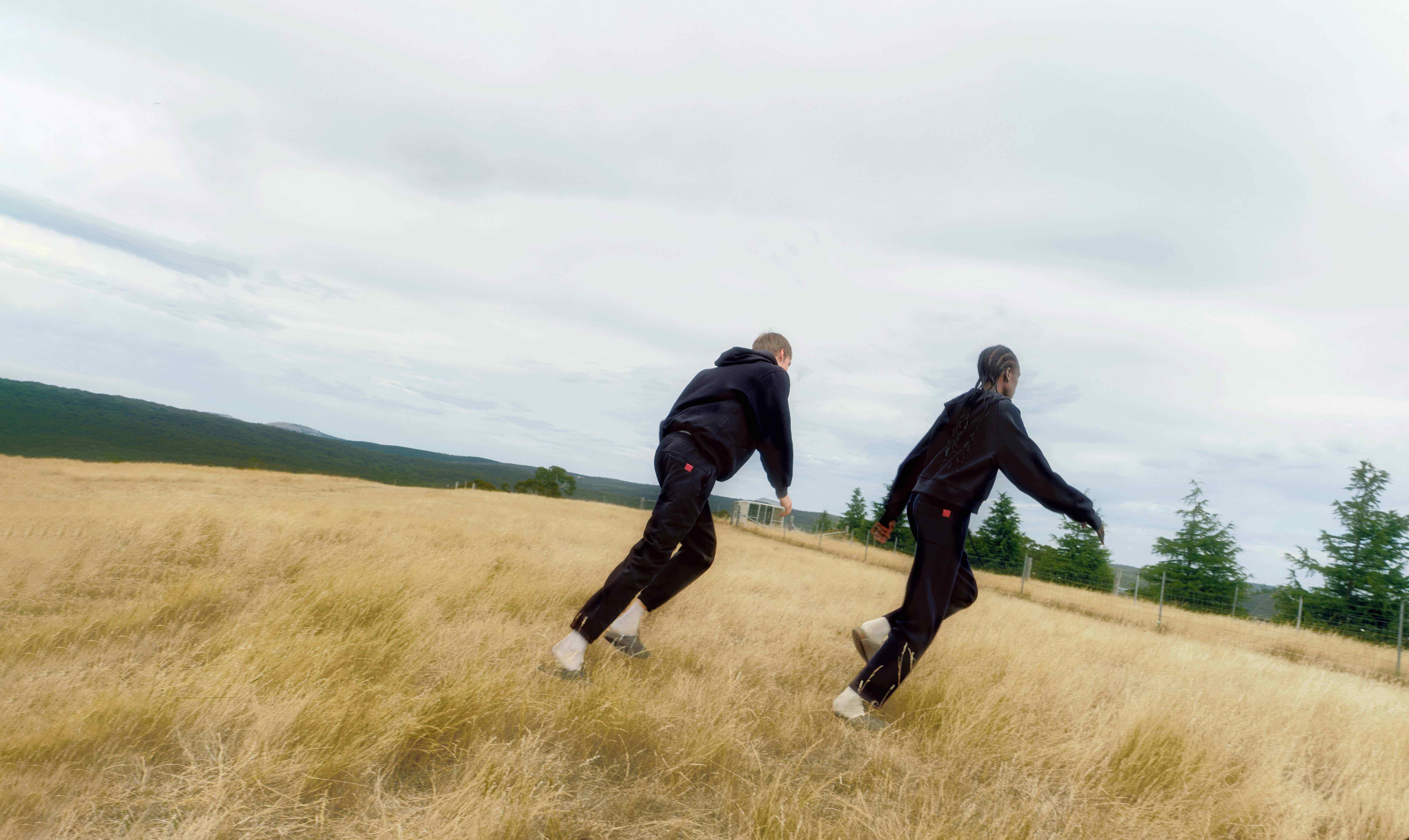 Two people in black outfits run through a grassy field under a cloudy sky.