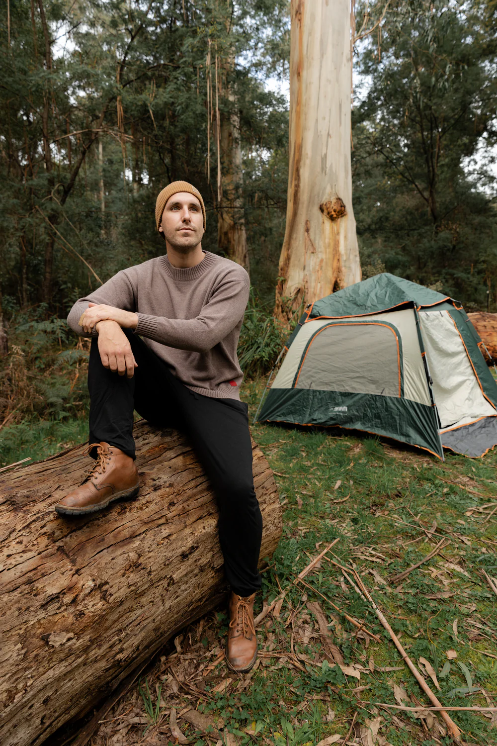 Man sitting on a log near a tent in a forest setting.