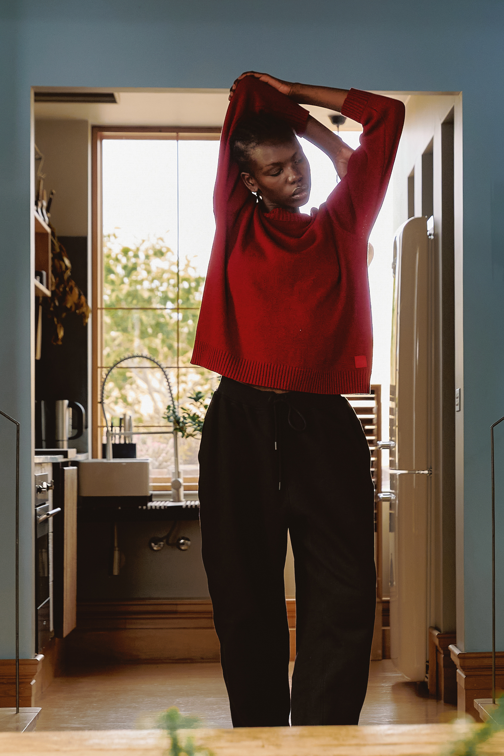 A person stretching in a kitchen, wearing a red sweater and black pants.