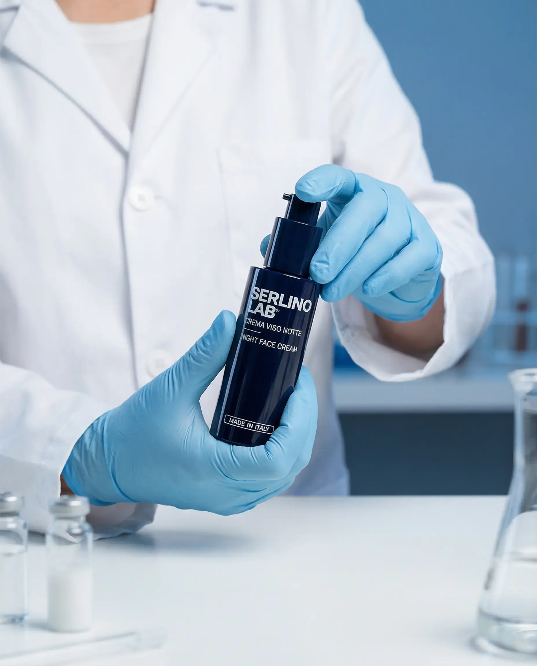 A person in a lab coat and blue gloves holds a bottle of Serlino Lab night face cream.