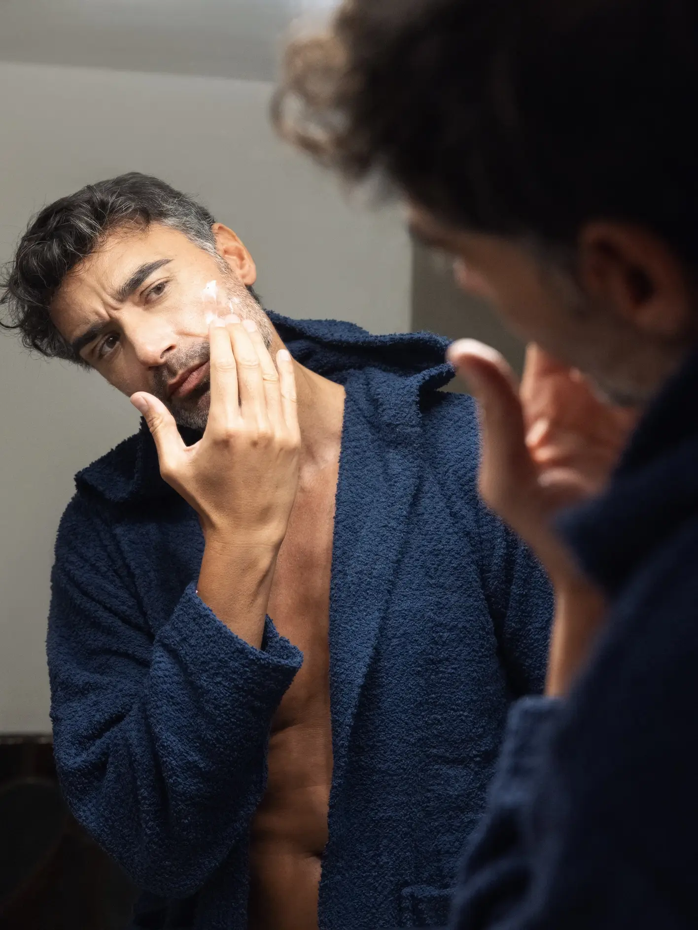 Man in blue robe applying cream to his face in the mirror.