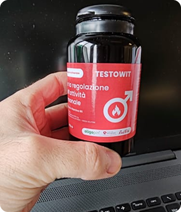 Hand holding a bottle with a red label in front of a laptop.