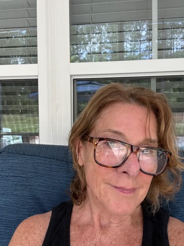 A woman with light brown hair and tortoiseshell glasses sits on a blue couch in front of windows.