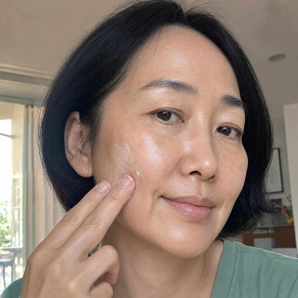 A woman with short black hair smiles while applying white cream to her cheek.