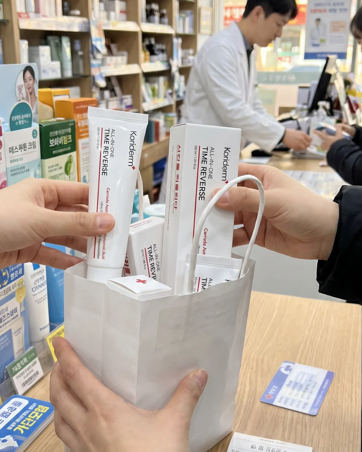 A person holds a white paper bag filled with Koriderm skincare products inside a pharmacy.