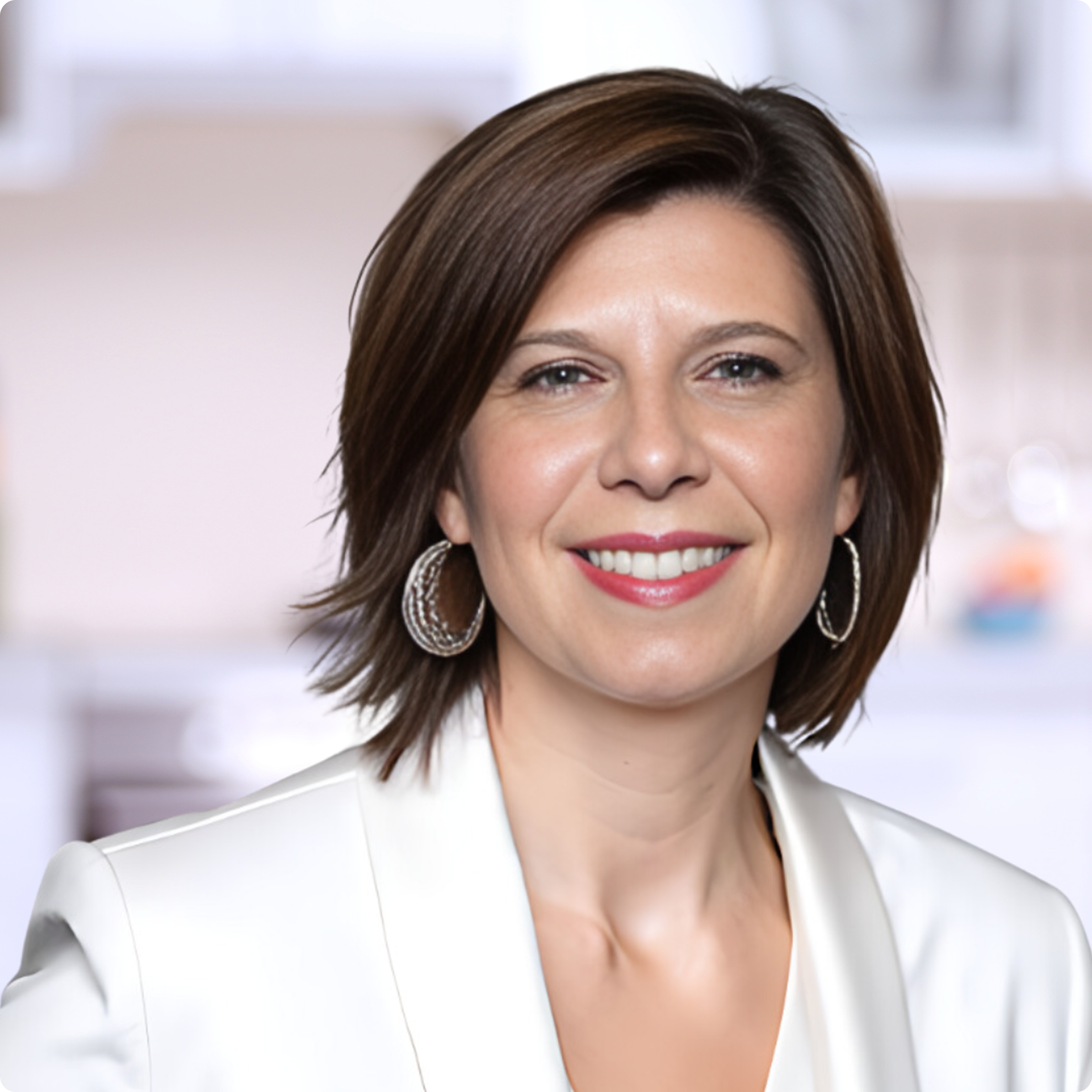 A headshot of a smiling woman with short brown hair, wearing a white blazer and silver hoop earrings.