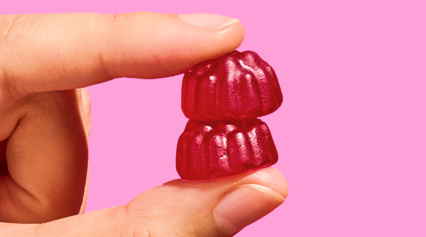 A close-up of a hand holding two stacked red gummies against a bright pink background.
