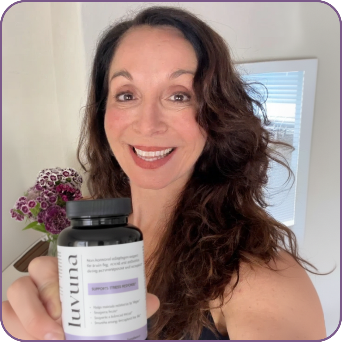 Smiling person holding a bottle of Luvuna supplement in front of flowers.
