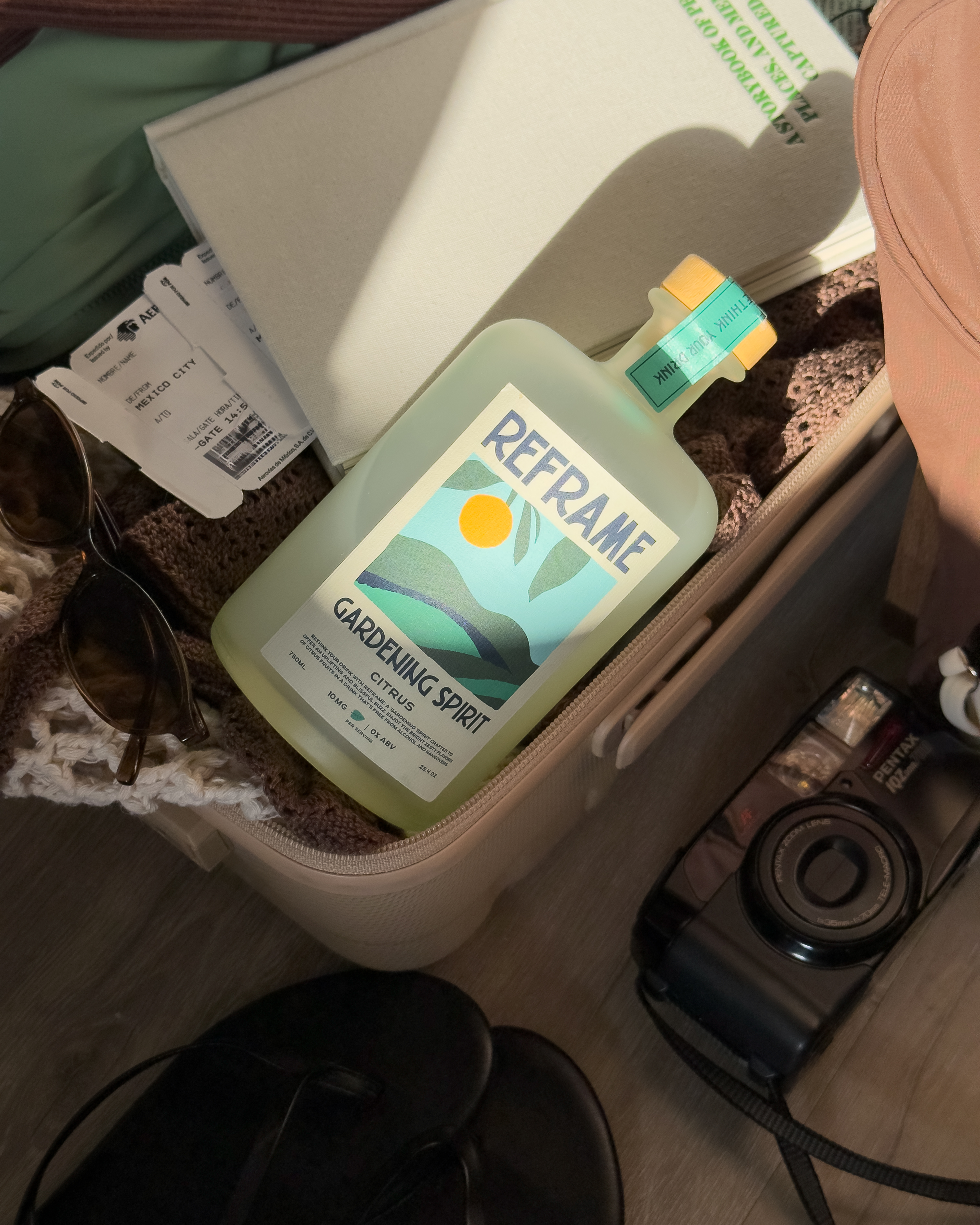 A bottle labeled 'Reframe Gardening Spirit' in a suitcase with sunglasses and a camera.