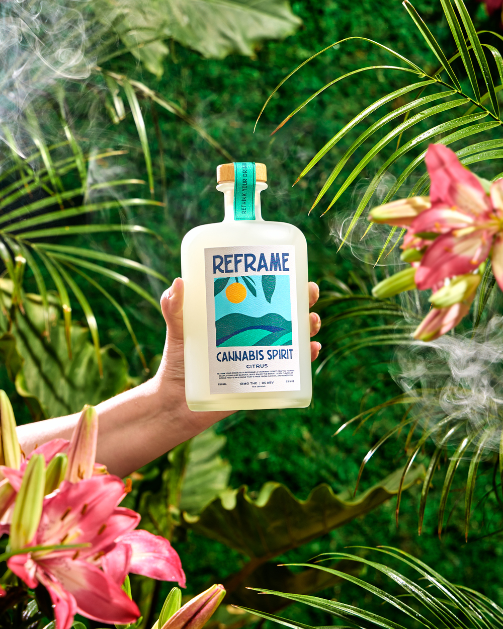 Hand holding a bottle labeled 'Reframe Cannabis Spirit' among tropical plants and flowers.