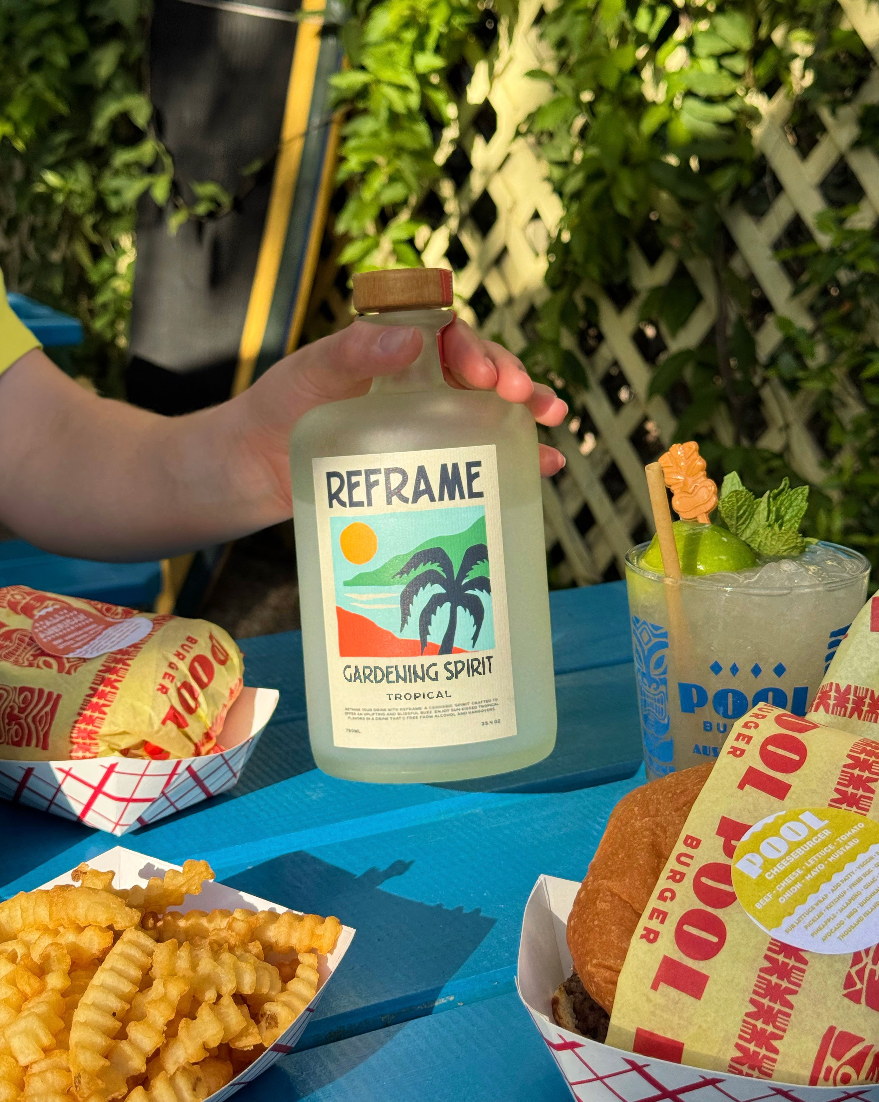 Hand holding a bottle labeled 'Reframe Gardening Spirit' near fries, burger, and drink on a blue table.