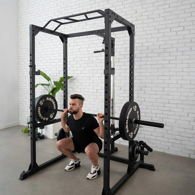 A man performs a weighted barbell squat inside a black power rack in front of a white brick wall.