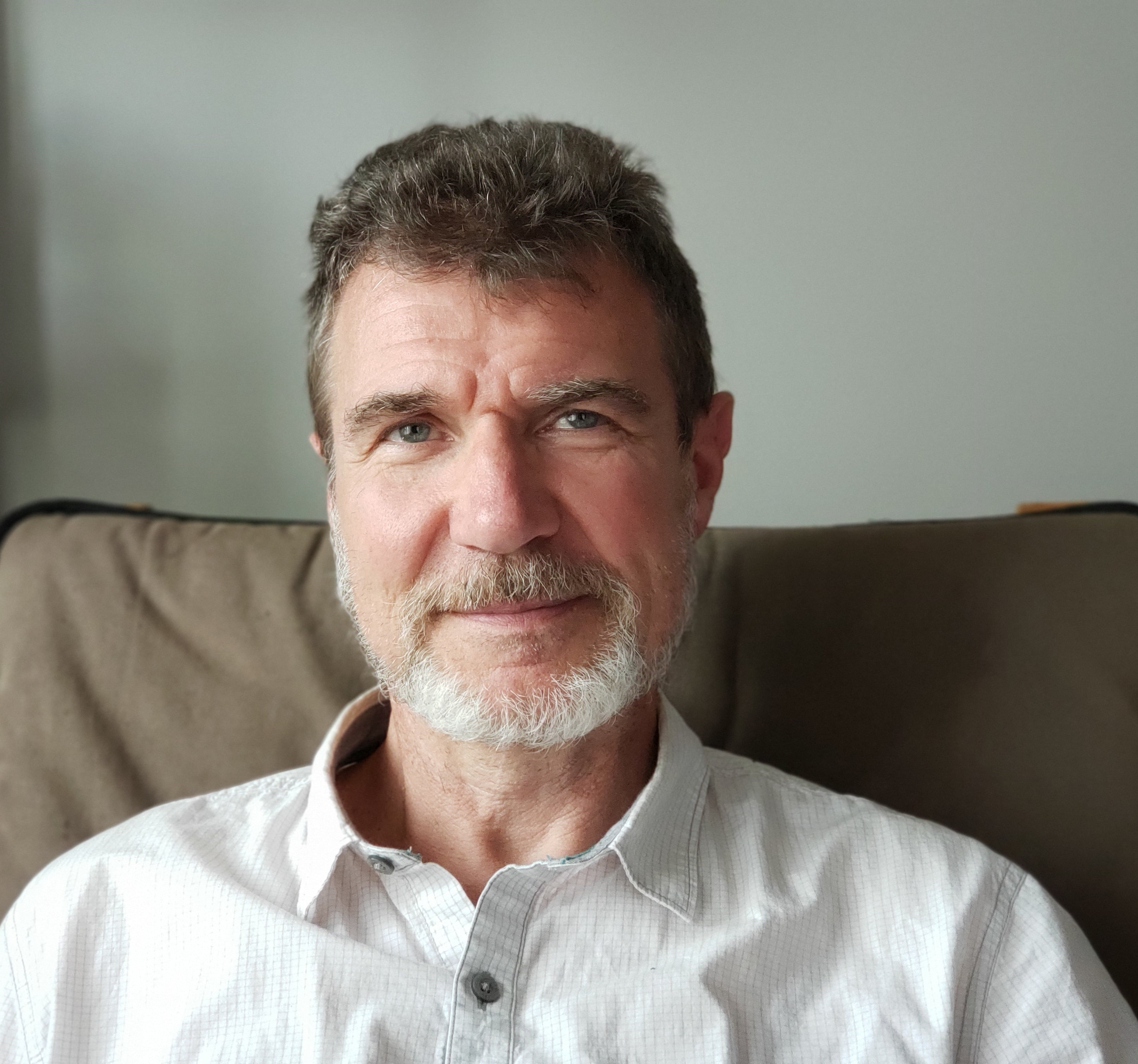 A man with a beard and mustache, wearing a light-colored shirt, sitting on a couch.