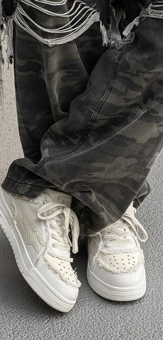 A close-up of a person wearing white distressed sneakers and dark camouflage pants on a gray surface.