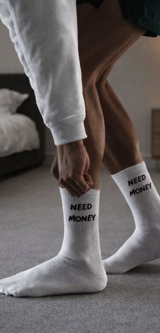 A person puts on white socks with the words 'NEED MONEY' printed on the side.
