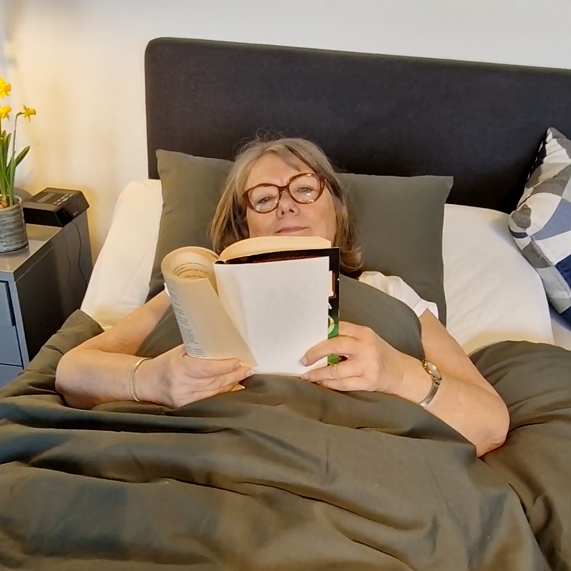 Person lying in bed reading a book, with glasses and gray bedding.
