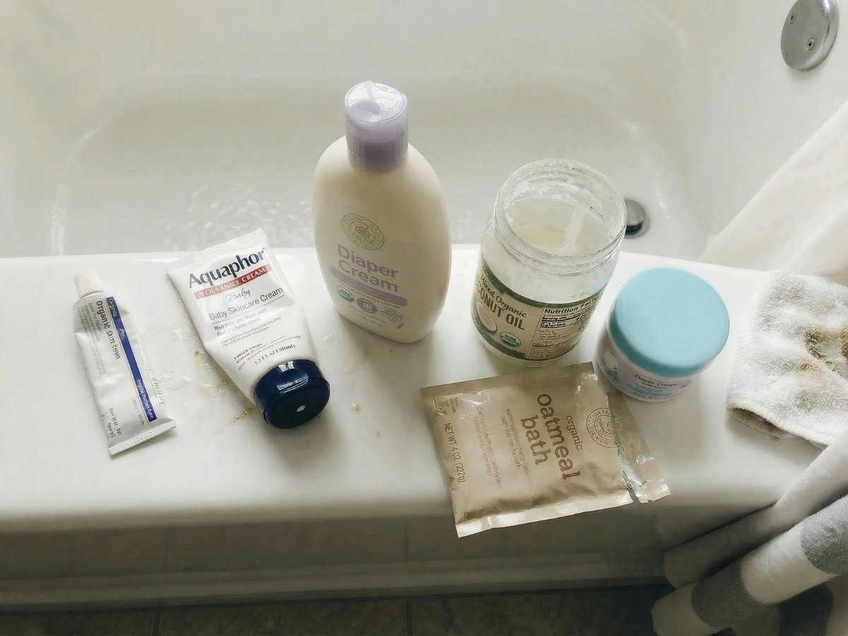 Various baby skincare products, including creams, oils, and an oatmeal bath packet, sit on a bathtub ledge.