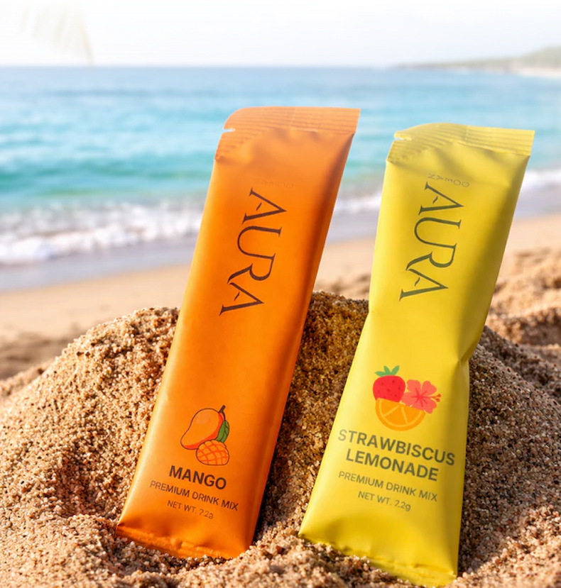 Two packets of Aura brand drink mix, mango and lemonade flavors, sitting in the sand on a beach.