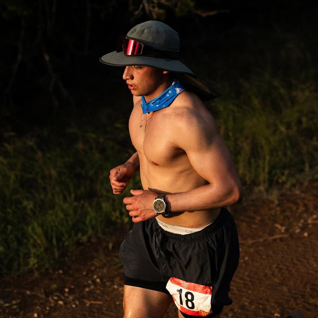 Shirtless runner with a hat and blue bandana, wearing a race bib number 18.